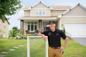 Window Companies Minocqua WI View of Renewal by Andersen employee wearing black polo and khakis leaning against a casement window while standing in front of two-story home.